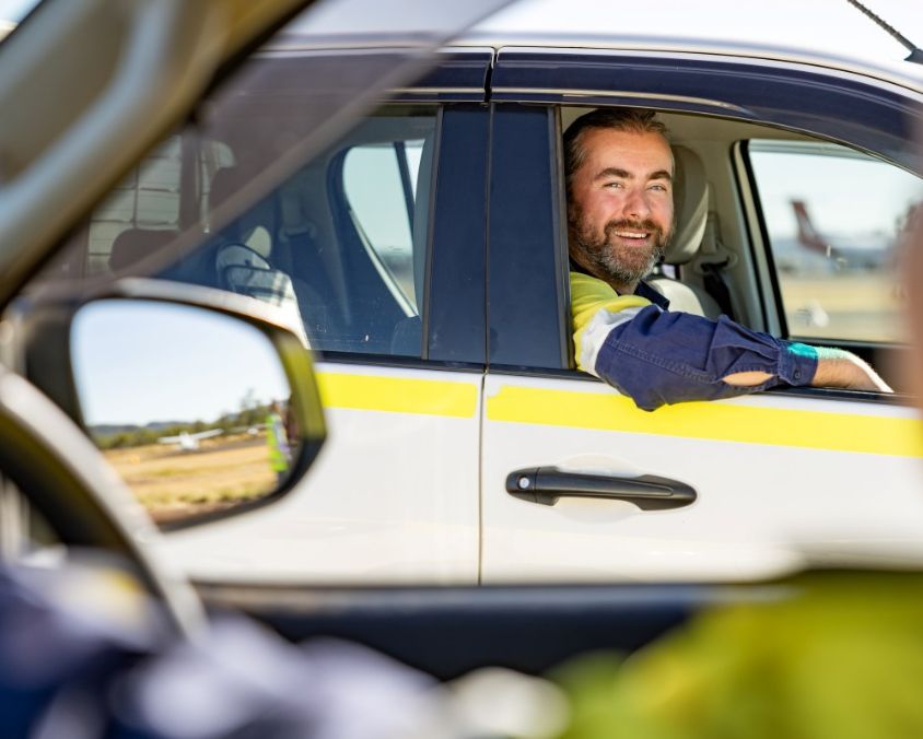 Person seated in the driver's seat of a white vehicle with a yellow stripe, interacting through the open window, airplanes visible in the background at an airfield or airport.
