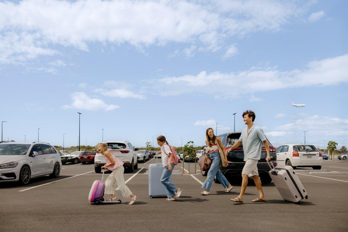 Family of four walking through an airport parking lot with luggage, under a clear sky with an aircraft visible overhead.