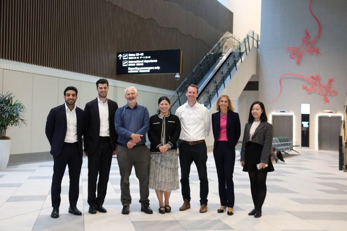 Group of business professionals and QAL investors standing inside Gold Coast Airport terminal near escalator and directional signage.