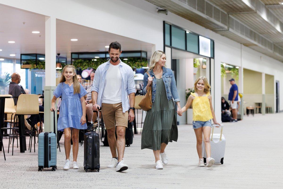 Family of four walking through Townsville Airport terminal with luggage, passing shops and seating areas.