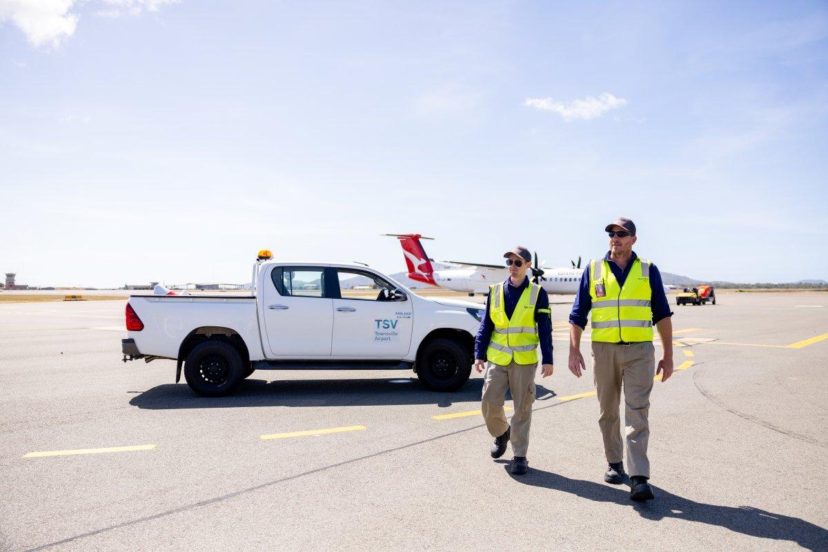 Airport operations staff walking on the tarmac near a utility vehicle and aircraft, representing roles requiring ASIC accreditation.