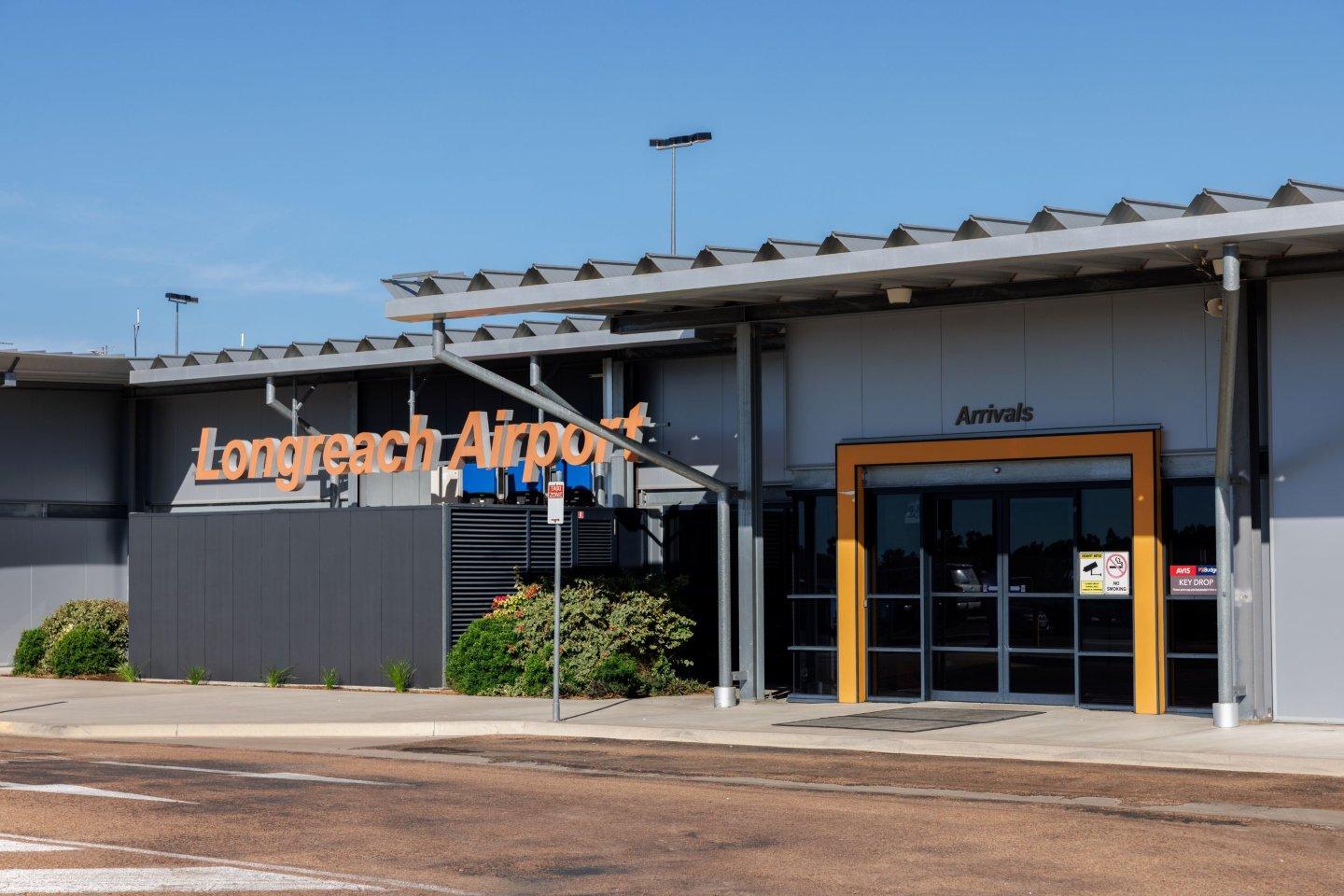 Exterior view of Longreach Airport entrance with signage for "Longreach Airport" and "Arrivals"; modern building with metal roof and greenery under a clear blue sky.