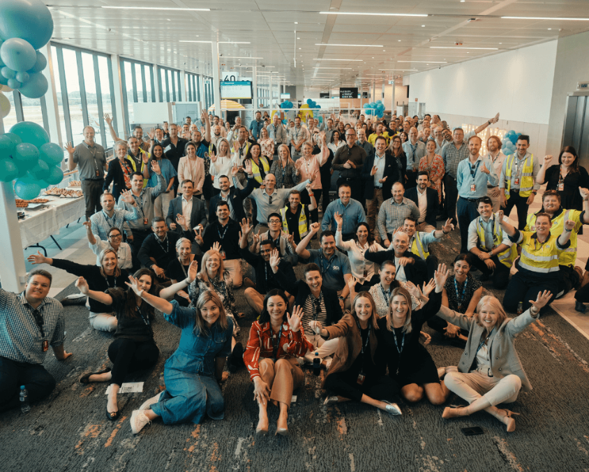 A large, cheerful group of airport staff and partners pose together inside a terminal at Gold Coast Airport, celebrating with balloons and snacks in a bright, festive atmosphere.