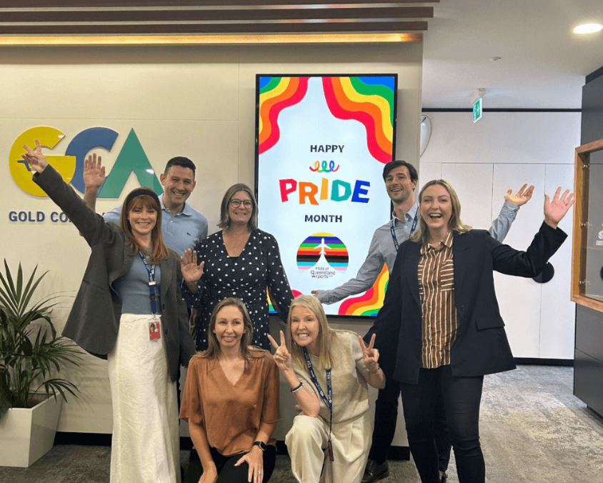 A group of Gold Coast Airport team members smile and pose enthusiastically in front of a digital sign that reads “Happy Pride Month,” featuring rainbow colours and inclusive messaging.