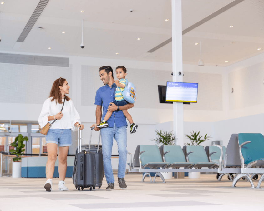 A young family walks through an airport terminal, with the father carrying a child and the mother pulling a suitcase. They appear happy and relaxed in a bright, modern waiting area.