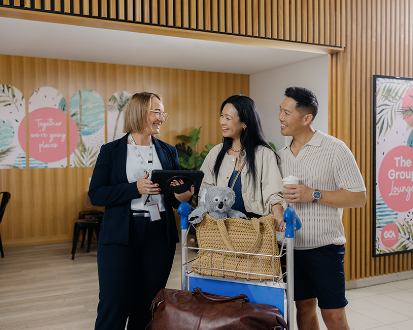 A smiling airport staff member assists two travellers with a luggage trolley in front of colourful signage for The Group Lounge at Gold Coast Airport. The couple looks happy and engaged in conversation.