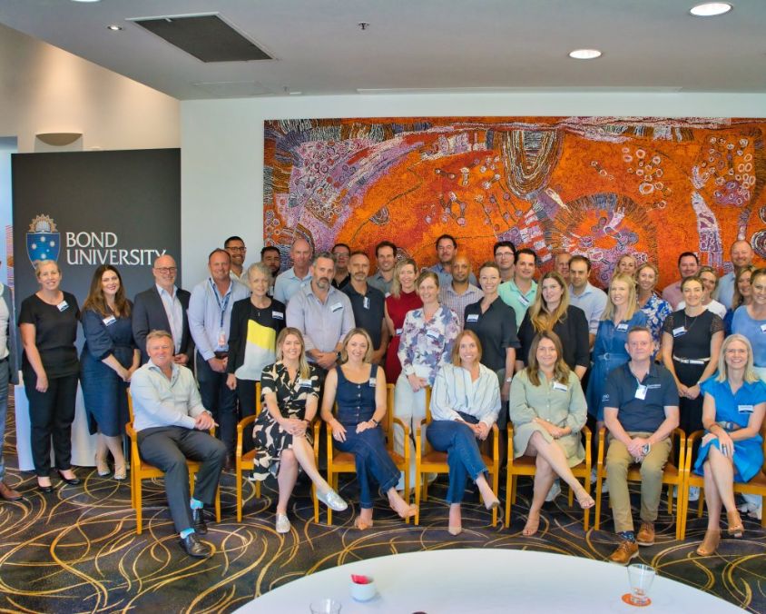 Group of people posing in front of a large orange and red artwork at a Bond University event; some seated in front, others standing behind; Bond University sign and logo visible on the left.
