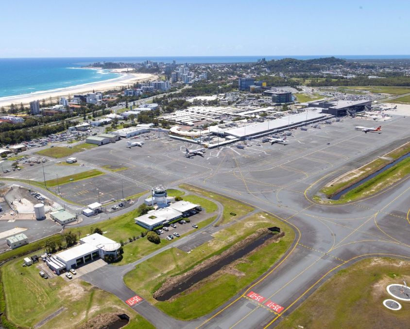 Aerial view of Gold Coast Airport near the coast, showing runways, terminals, and parked aircraft with city and beach in the background.