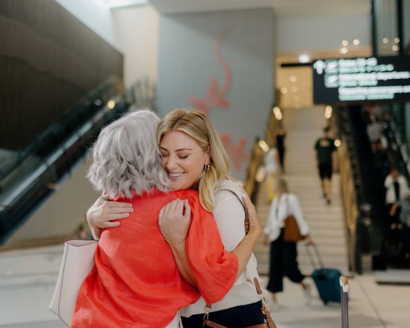 Two people embracing at the Gold Coast Airport terminal near escalators and a staircase, with signage and people in the background