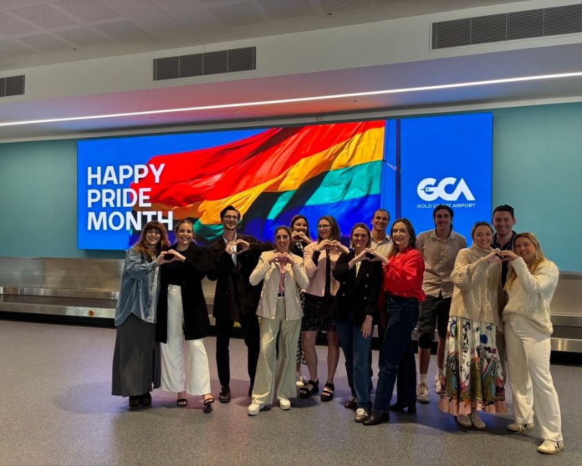 Group of people standing in front of a large digital display at Gold Coast Airport featuring a rainbow flag and the message “Happy Pride Month.” The group poses in two rows, making heart shapes with their hands to celebrate LGBTQ+ inclusion.