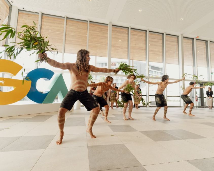 Traditional dance performance inside a GCA's International Arrivals Hall, with dancers holding green foliage and wearing body paint. A large “GCA” sign in yellow, blue, and green is visible in the background, with observers watching the ceremony.