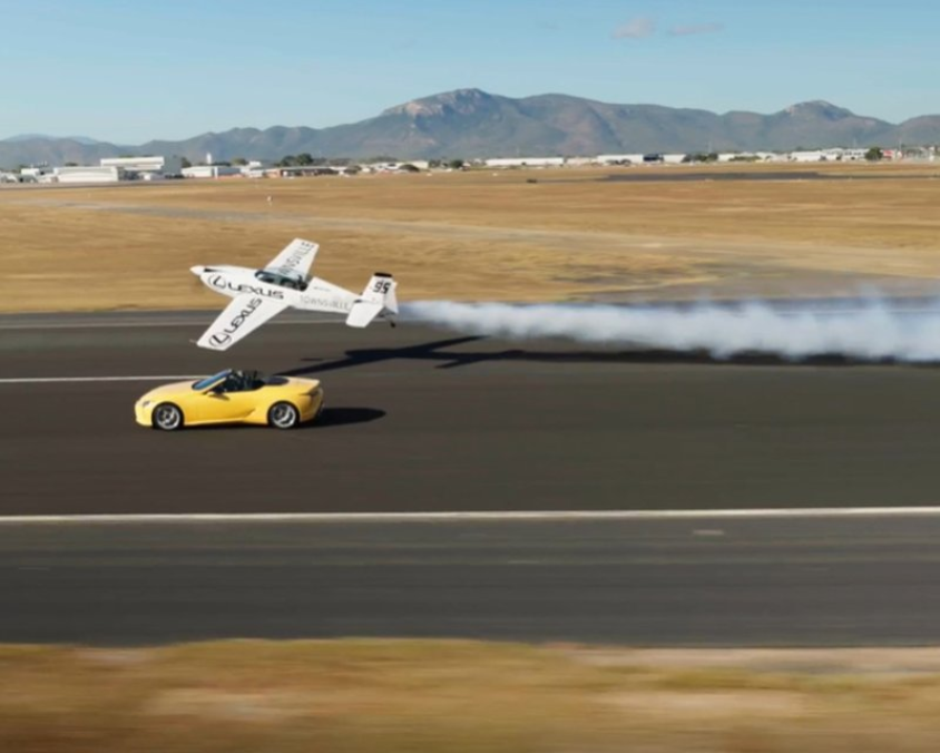 Yellow sports car driving on a runway at Townsville Airport with a low-flying white airplane trailing smoke overhead, and airfield buildings and mountains in the background.
