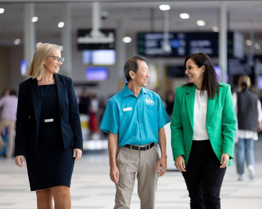 Three professionally dressed individuals walk through a busy airport terminal, smiling and engaged in conversation. Flight information boards and passengers are visible in the background.