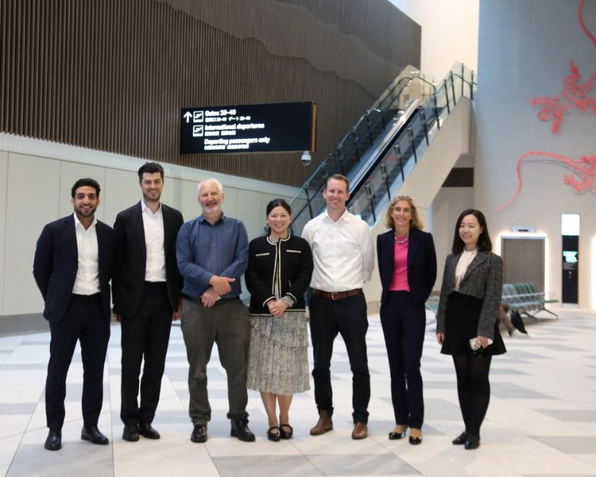 Group of business professionals and QAL investors standing inside Gold Coast Airport terminal near escalator and directional signage.