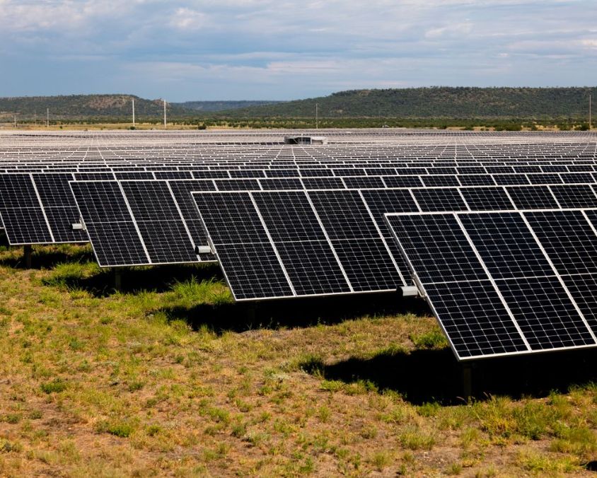Wide view of a solar panel farm with rows of panels on grassy terrain, set against hills and a partly cloudy sky.
